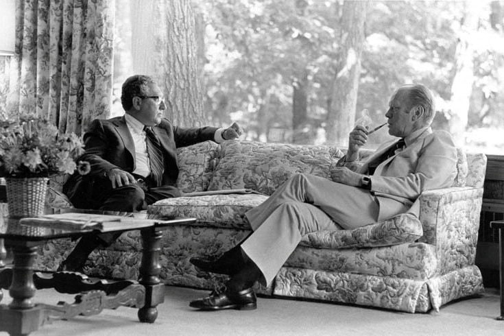 A black-and-white photo of two men sitting on opposite ends of a plush couch, one smoking a pipe.