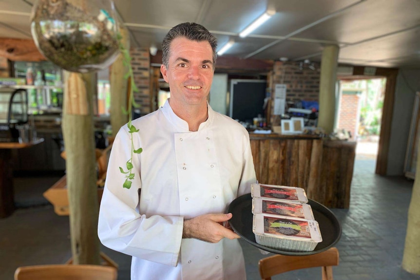 Chef stands in a room holding three takeaway meal boxes on a round black tray.