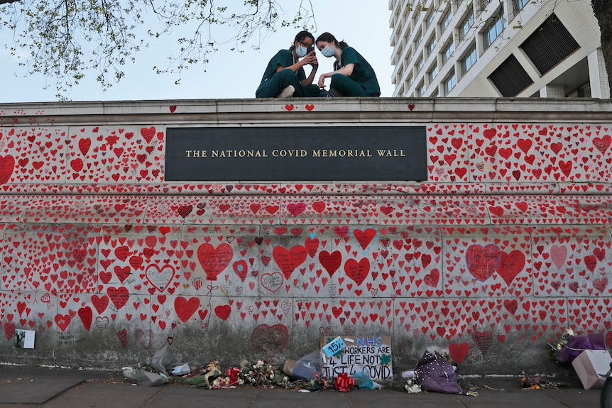 Two masked healthworkers sit atop a memorial wall covered in stylised hearts drawn in tribute to those who died from COVID.