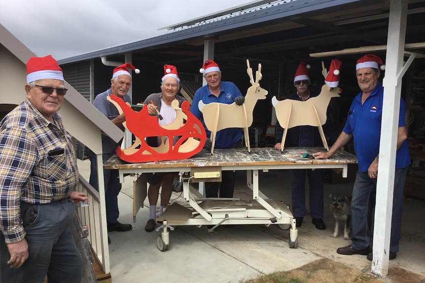 Group of older men wearing Santa hats crowd around a table that has a reindeer display.