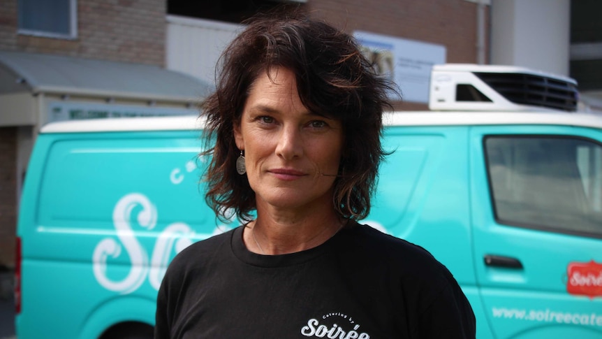 A woman in a black shirt stands in front of a blue van