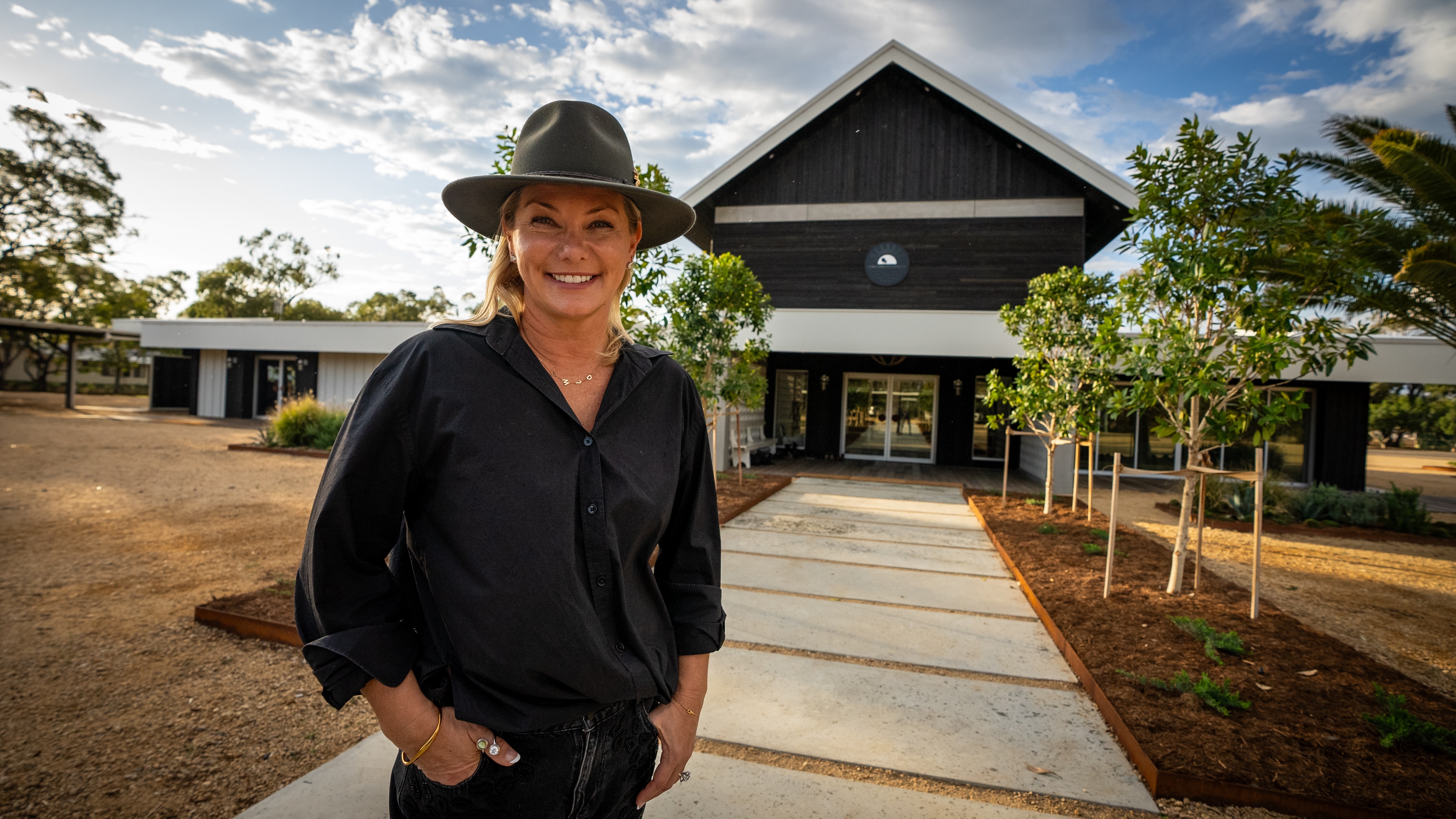 A woman in hat, hands in pocket, stands smiling on the path to her country house.