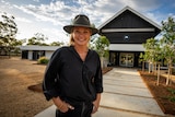 A woman stands, smiling, hands in pocket on the path to her house. 