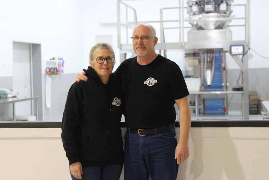 A man and a woman stand with their arms around each other, a stainless steel machine behind them