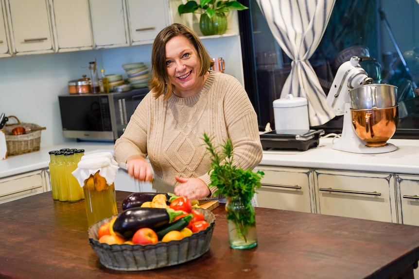 A woman in a brown knitted sweater stands over a rustic kitchen bench cutting capsicums, smiling.