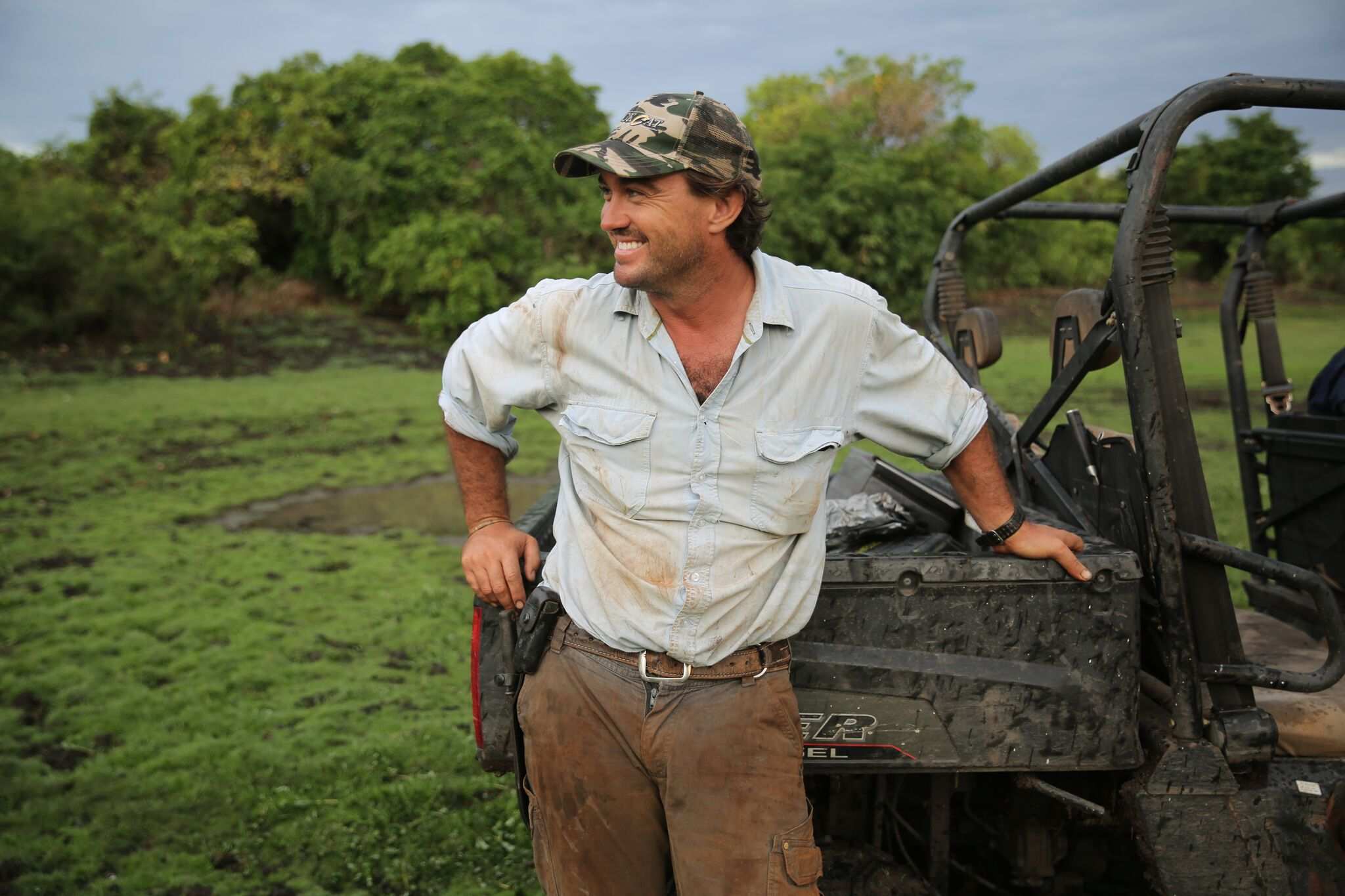 A white man in a khaki long sleeve shirt, brown pants, leaning up against a buggy in green land.