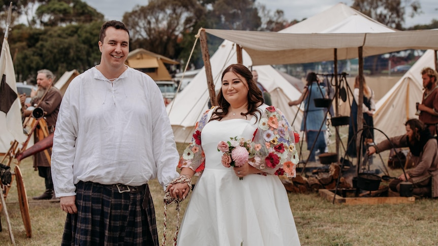 A groom and a bride holding hands with a rope tied around their hands, and medieval camp in the background