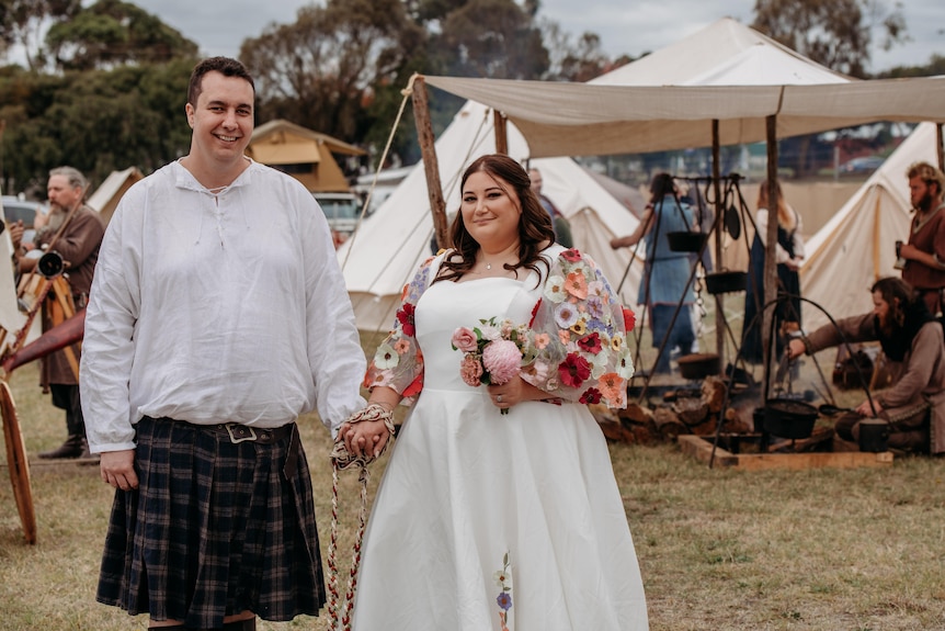 A groom and a bride holding hands with a rope tied around their hands, and medieval camp in the background