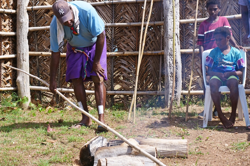 man with blue shirt bows down with long bamboo stick. Children watch on in background. There is smoke coming from front of stick