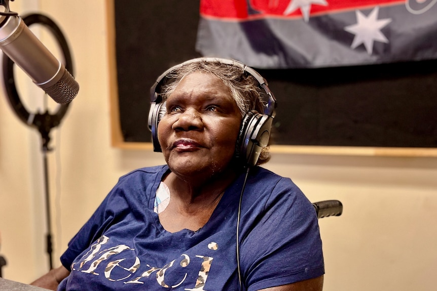 An old Indigenous woman sits with headphones on and a microphone in front of her. 