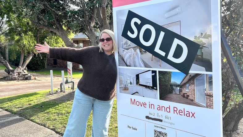 A blonde woman in sunglasses stands in front of a "sold" house sign