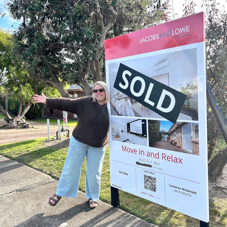 A blonde woman in sunglasses stands in front of a "sold" house sign