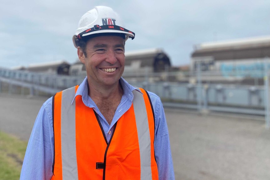 A man wearing a hard hat and hi-vis vest smiling in front of a row of train wagons and a grain conveyor.