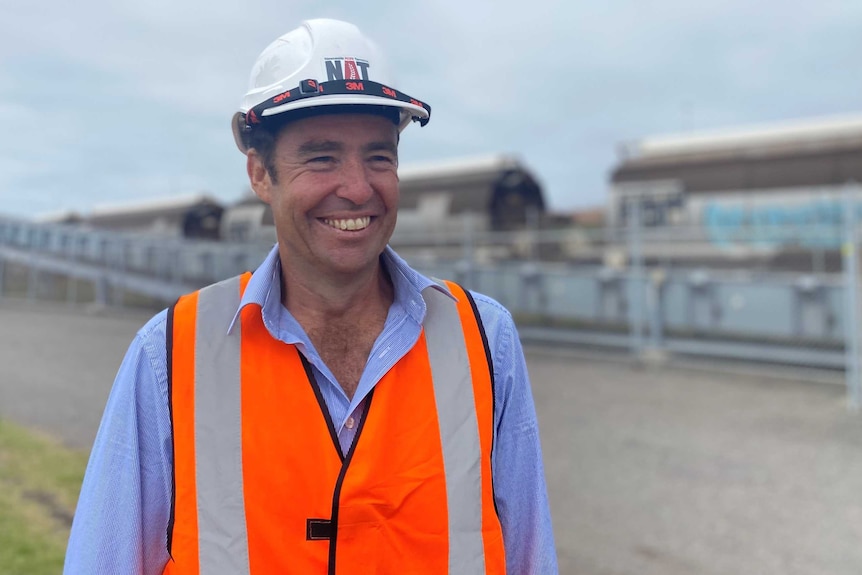 A man wearing a hard hat and hi-vis vest smiling in front of a row of train wagons and a grain conveyor.