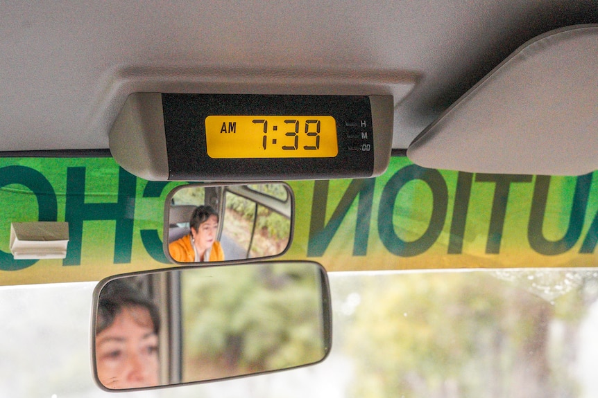 A woman with dark hair wearing a yellow jumper is reflected in a rear view mirror of a mini bus.