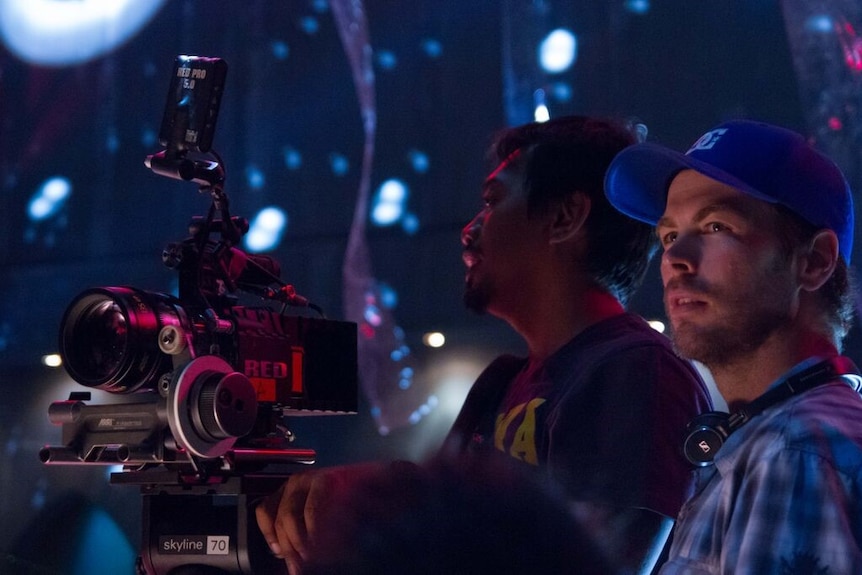 Corey Pearson stands in a dark room with another man operating a camera.