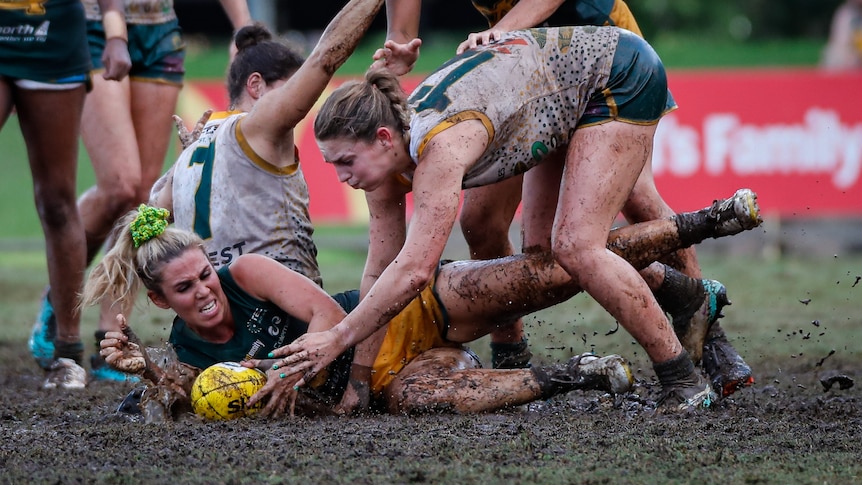 two women fight for a football on a muddy oval.