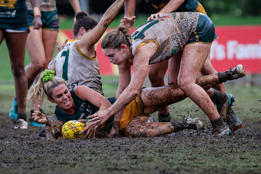 two women fight for a football on a muddy oval.