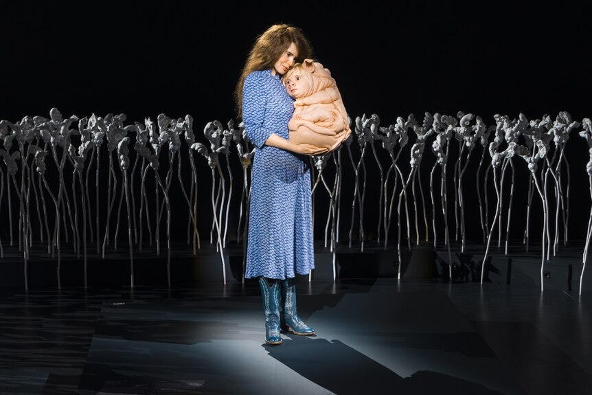 Long-haired woman in blue dress holding a genetically-engineered baby with pig features, in field of white sculpture 'flowers'.