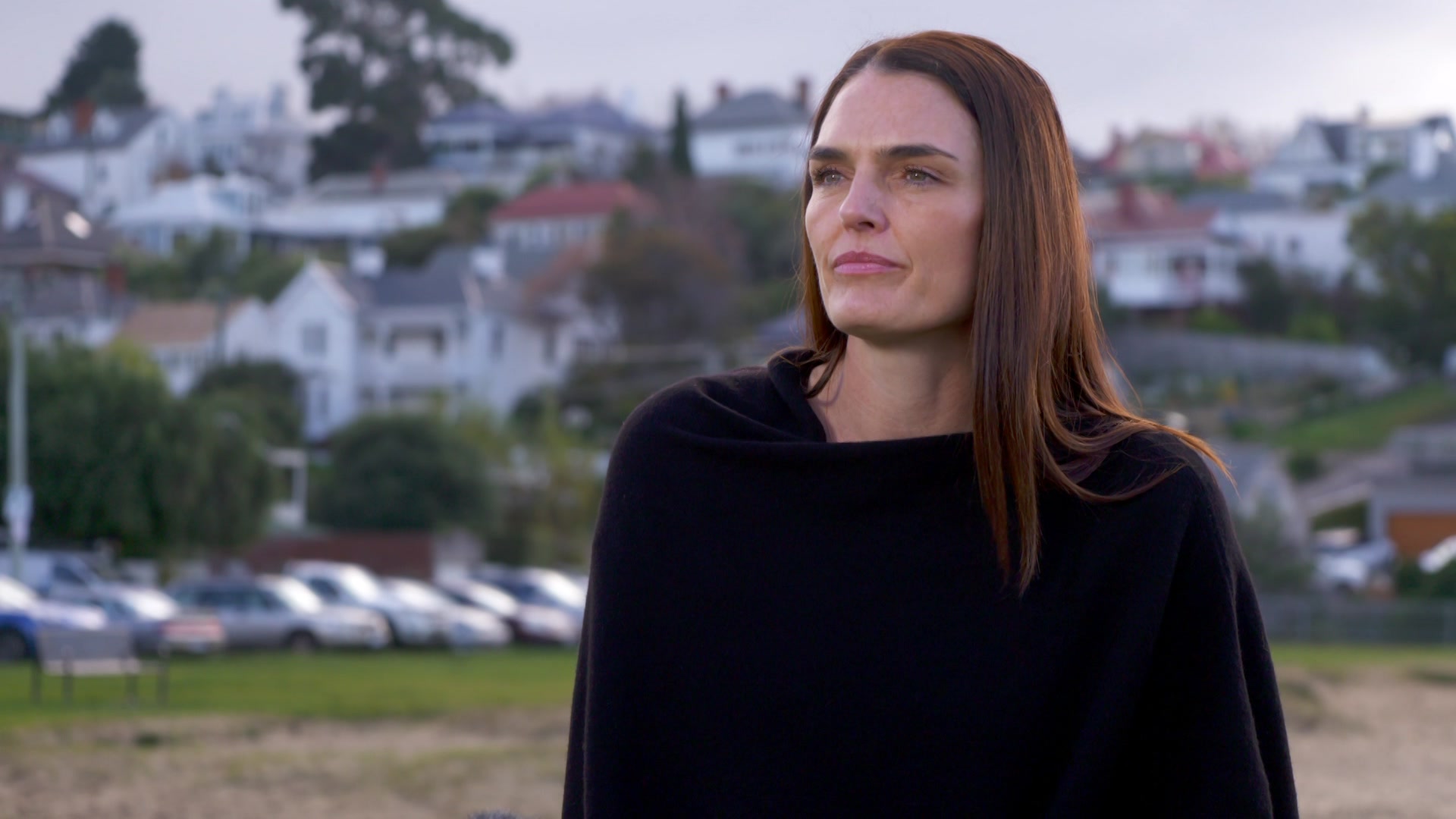 Tania Hunt wears a black jumper, standing in a park with houses on hill behind her