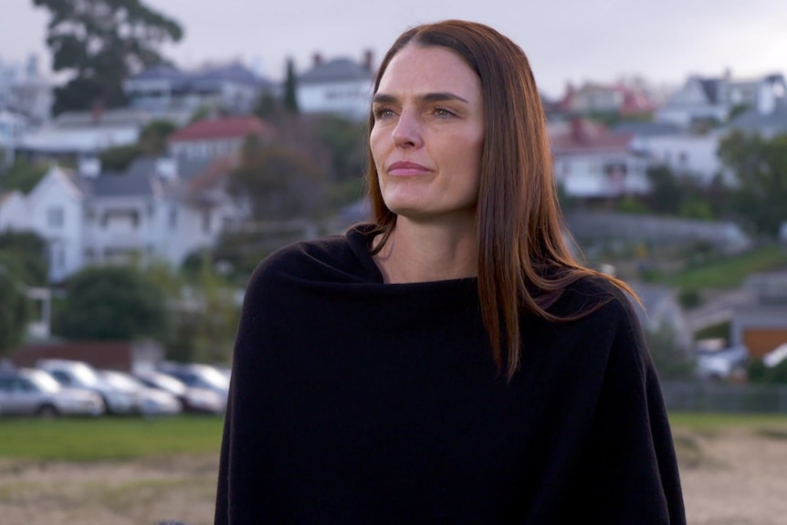Tania Hunt wears a black jumper, standing in a park with houses on hill behind her