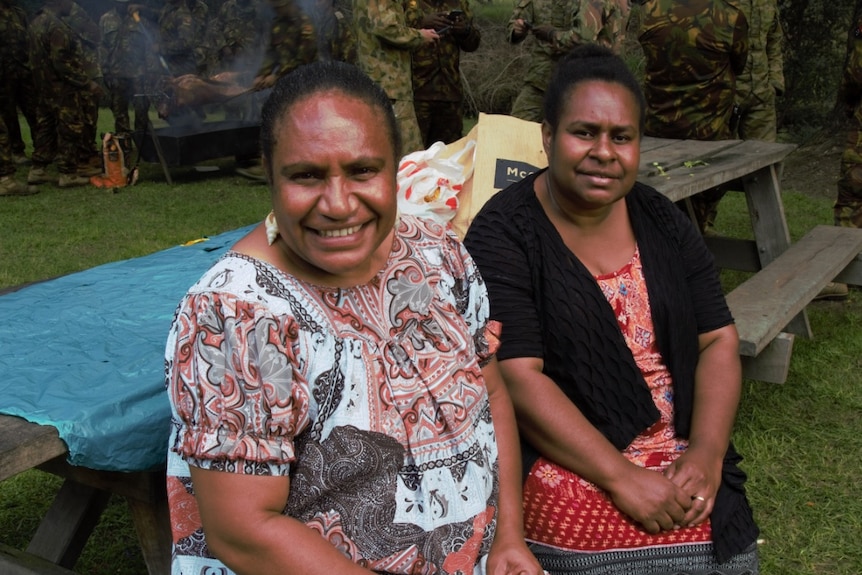 Two women in colourful patterned red shirts sit on a wooden bench with army troops mingling the background.