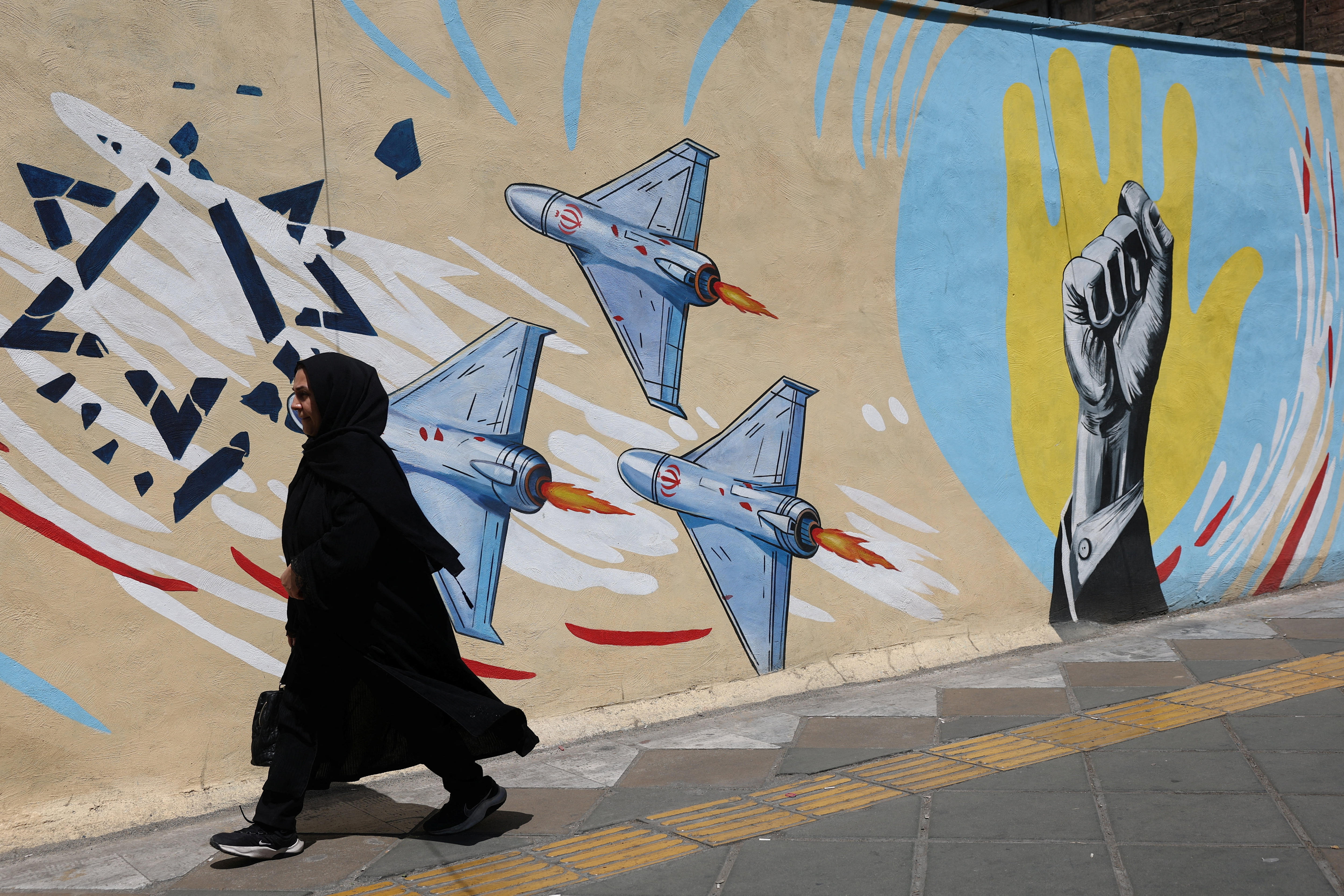 A woman in burqa walks on a street in Tehran infront of wall with IRGC planes on it.