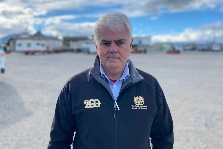 A grey-haired man stands on a showground in front of a line of campervans.