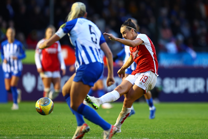 A player dressed in red and white kicks past a player in blue and white in a women's soccer match.