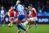 A player dressed in red and white kicks past a player in blue and white in a women's soccer match.