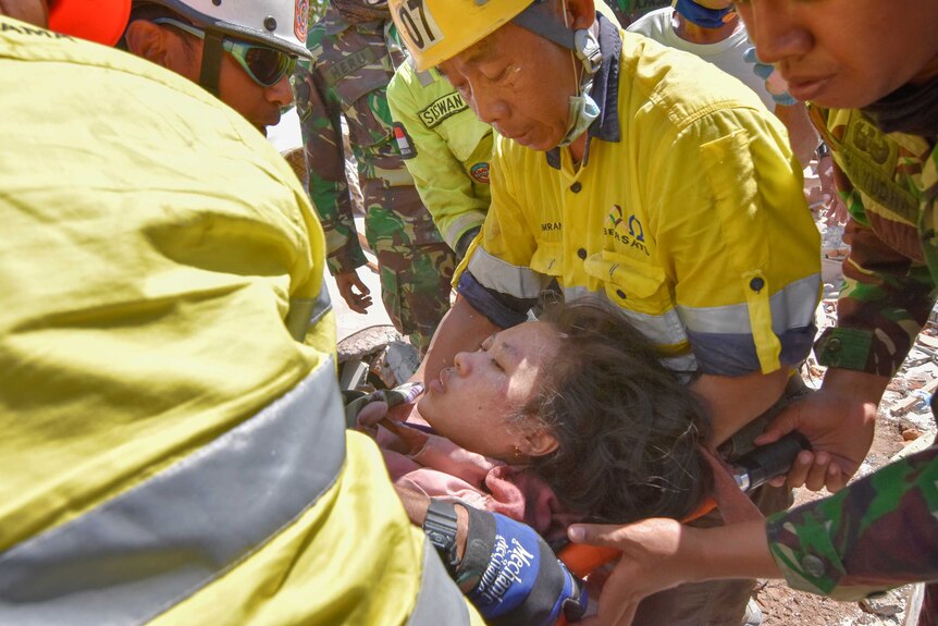 A tight shot shows people dressed in high-vis and military workwear attend to a woman with her eyes closed on a stretcher