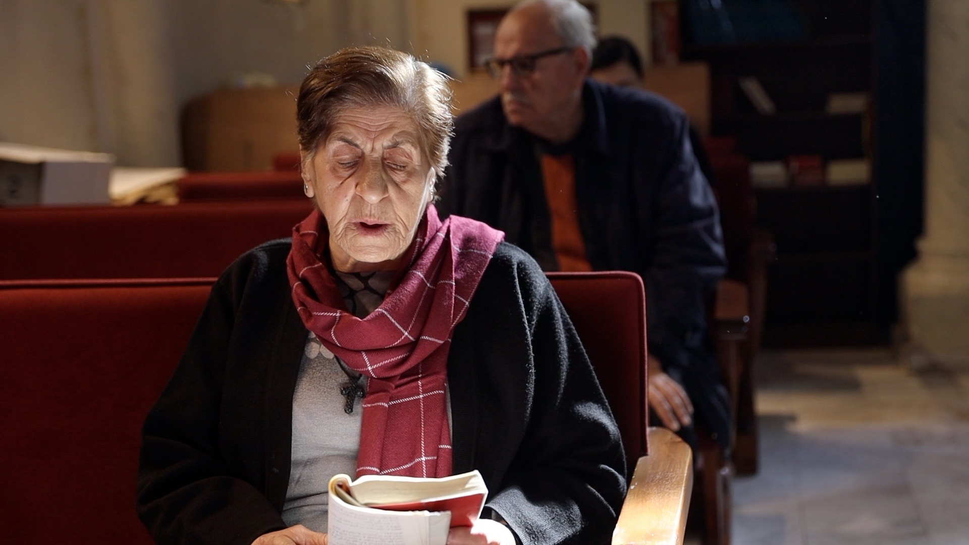 Una anciana sentada en el banco de una iglesia con una cruz y un pañuelo alrededor del cuello, sosteniendo un libro abierto mientras una luz brillante brilla sobre ella
