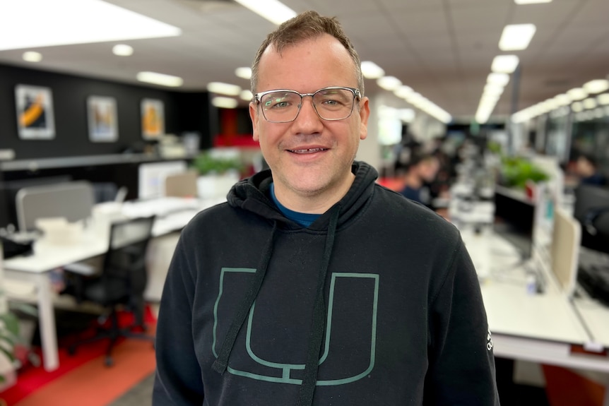 A man with glasses, a receding hairline and wearing a black hoodie stands in an office and smiles to the camera.