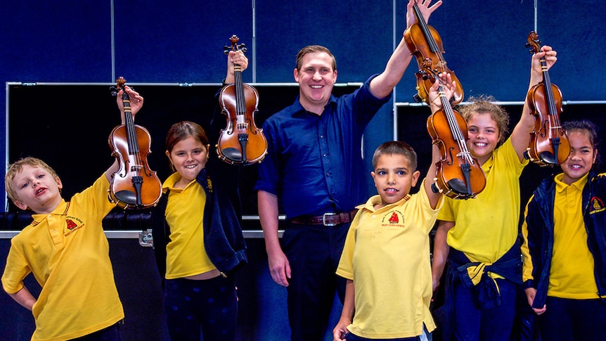 Five primary students and a teacher smile and hold up violins in front of a blue background.