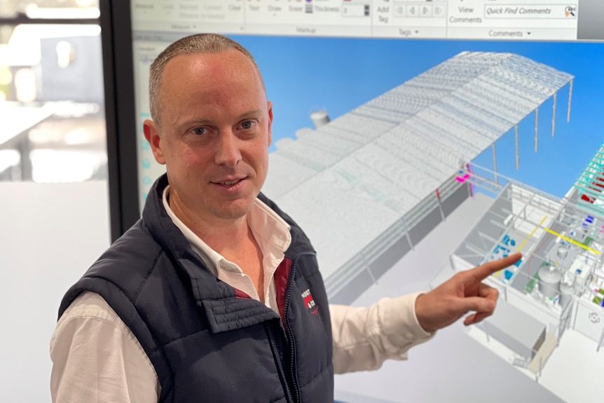medium shot of a man pointing to a factory layout image on a screen behind him