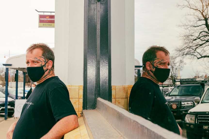 Brian Warton leans against a pub window, wearing a face mask, and his reflection behind him.