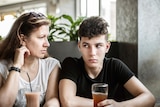 A mum and son sit in a cafe, the mum looks at her kid with concern, the son looks annoyed