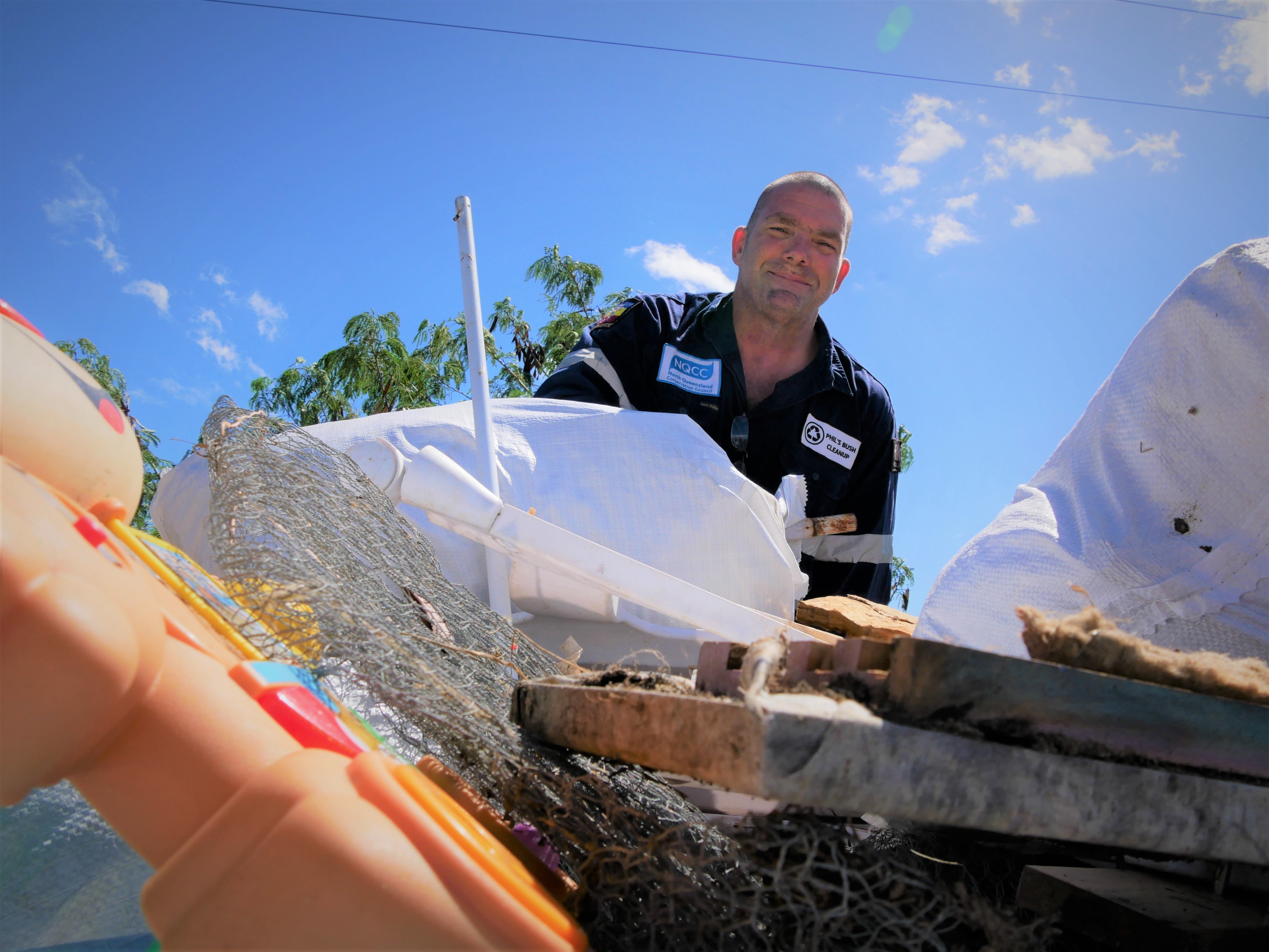 A man with shaved head and work shirt smiles down at camera above pile of rubbish. 