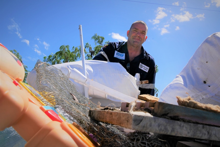 A man with shaved head and work shirt smiles down at camera above pile of rubbish. 