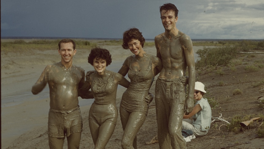 Four young people covered in mud posing for a photo next to a tidal flat.