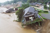 A house balances precariously on a cliff caused by erosion, as floodwaters flow underneath.