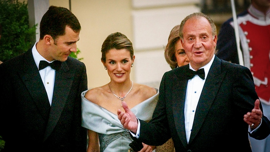 King Juan Carlos, an older man in a tuxedo, emphasises a point with his hands next to other opulently dressed people 
