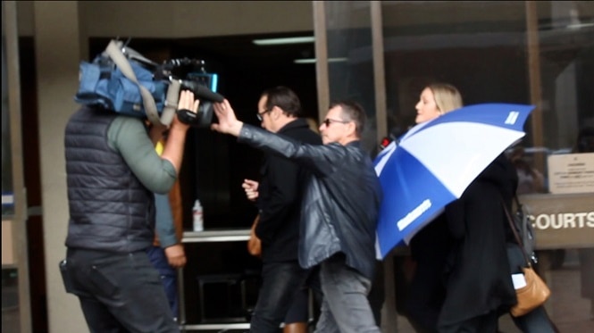 Katrina East arriving at Bunbury Court, hiding under an umbrella, while her father pushes away a camera man.