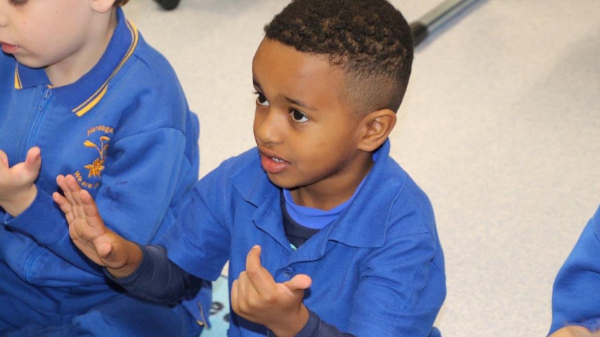 A young boy sits on the floor counting with his hands wearing a blue school uniform in class.