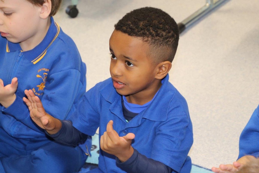 A young boy sits on the floor counting with his hands wearing a blue school uniform in class.