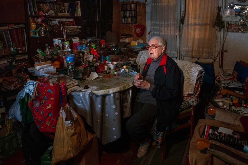 A woman sits alone in a dark, cluttered dining room.