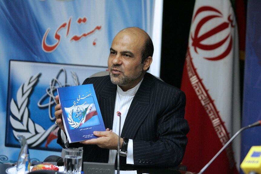 Man holding book with Iranian flag in background.