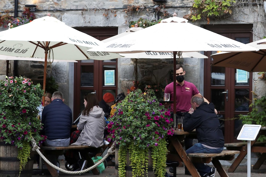 People drink in a Scottish beer garden, a barkeep collects glasses, while wearing a mask.