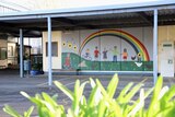 A mural with people painted under a rainbow holding hands sits under a shelter on school grounds.
