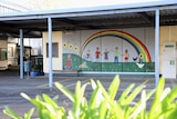 A mural with people painted under a rainbow holding hands sits under a shelter on school grounds.